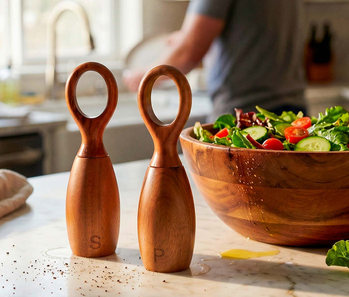 Wooden salt and pepper shakers on a kitchen counter with a salad in the background.
