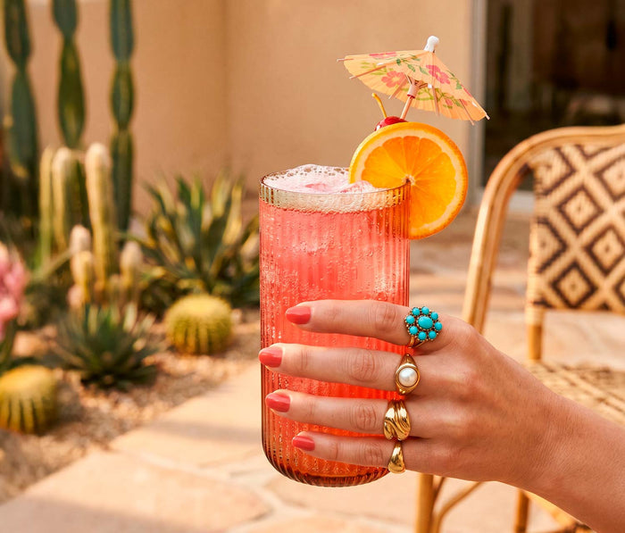 Hand holding a pink cocktail with an orange slice and umbrella, set against a desert-themed background.