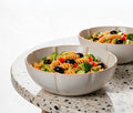 Two scalloped ceramic bowls filled with pasta salad, placed on a speckled stone table against a clean white background - Lollygag