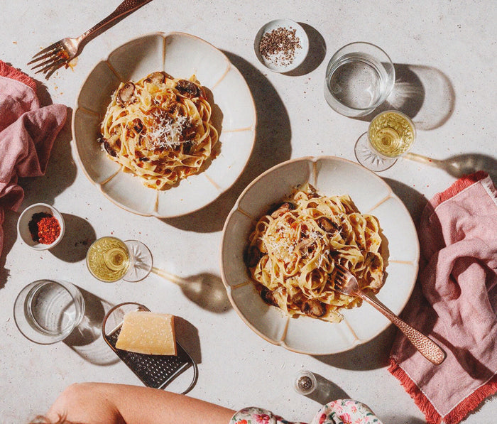 Two flower bowls of pasta served on large beige plates, arranged on a light stone surface with dining props including glasses of white wine, a grater, cheese, and pink napkins - Lollygag