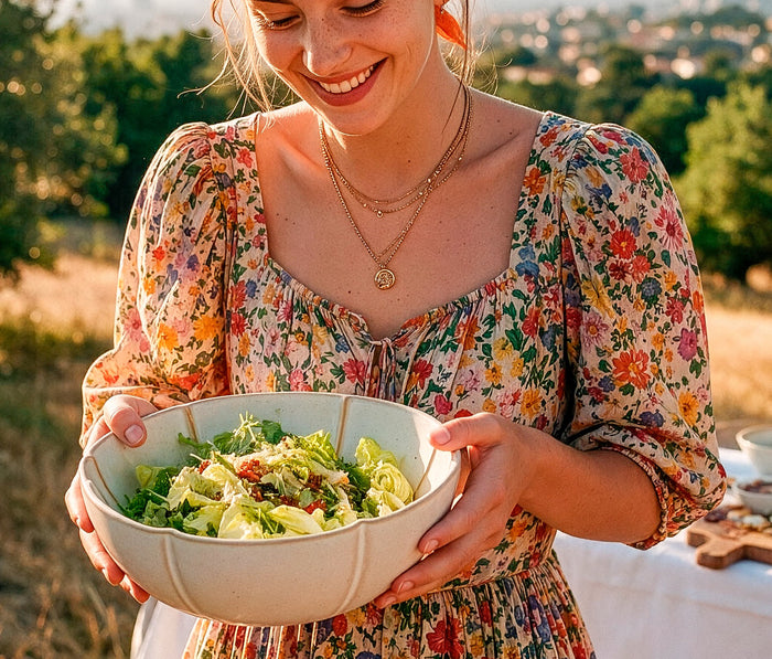 A woman outdoors wearing a colorful floral dress and holding a large earthtone- bowl filled with a green salad- Lollygag