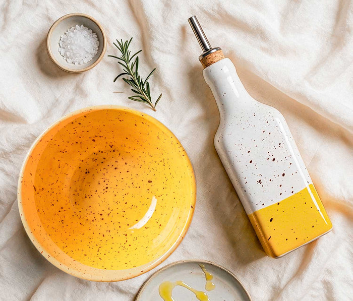 A ceramic oil bottle with a cork stopper and metal pour spout, painted white on top and bright yellow on the bottom, styled on a cream fabric surface beside a matching yellow bowl - Lollygag
