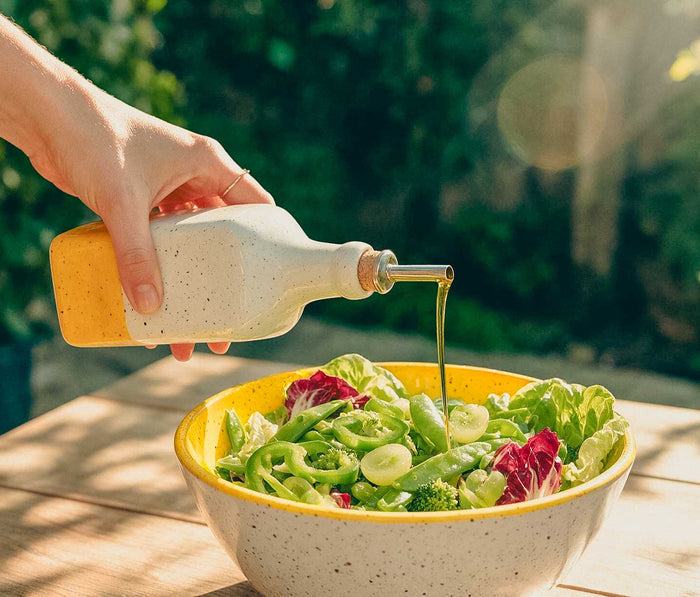 A hand holds a speckled white and yellow bottle, pouring olive oil over a fresh green salad in a matching speckled bowl on a wooden table outdoors - Lollygag