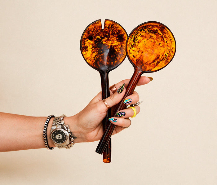 A hand holding two decorative serving spoons with amber and dark brown marbled handles and bowls, set against a plain beige background.