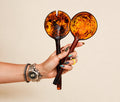 A hand holding two decorative serving spoons with amber and dark brown marbled handles and bowls, set against a plain beige background.