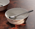 A ceramic bowl with a warm beige exterior featuring embossed floral and bird motifs, a white interior, and a silver spoon resting inside, set on a dark brown wooden surface.