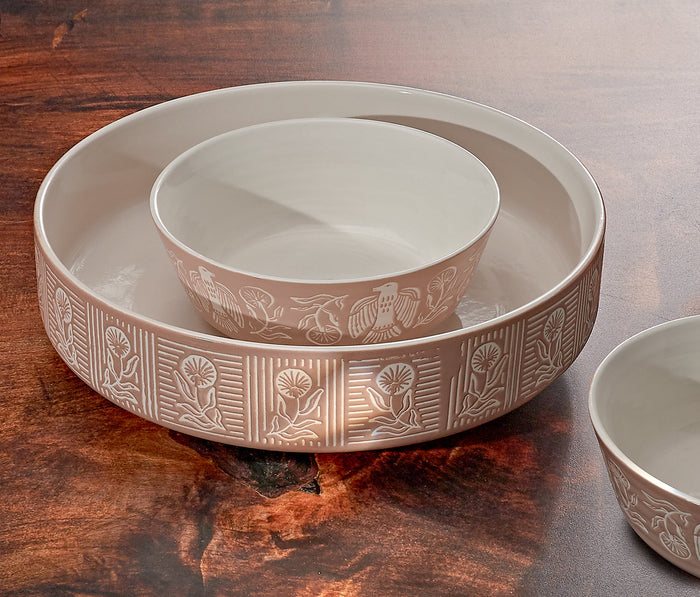 cream-colored bowl nested inside a larger shallow bowl, both with a light beige band decorated with embossed floral and geometric patterns, set on a dark wooden surface.