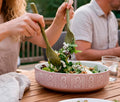 A pink ceramic salad bowl with a white floral pattern sits on a wooden table, being mixed with green leafy salad and crumbled cheese by two people at an outdoor dining table.