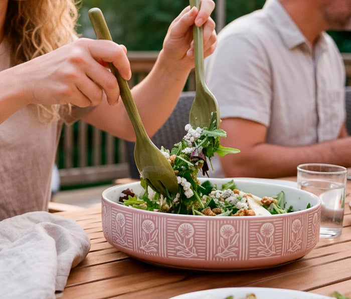 A pink ceramic salad bowl with a white floral pattern sits on a wooden table, being mixed with green leafy salad and crumbled cheese by two people at an outdoor dining table.