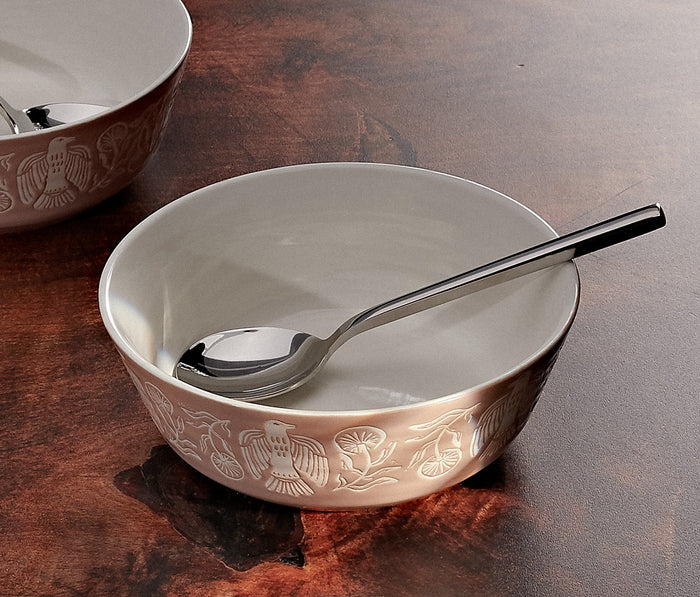 A ceramic bowl with a warm beige exterior featuring embossed floral and bird motifs, a white interior, and a silver spoon resting inside, set on a dark brown wooden surface.