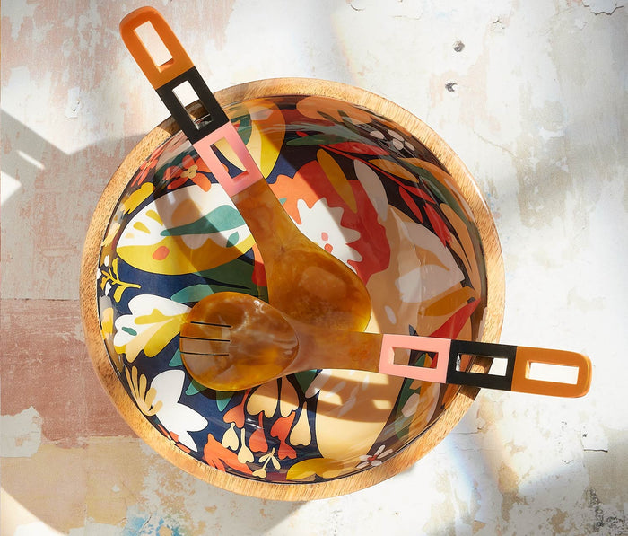 A top-down view of a round wooden bowl with a colorful floral pattern inside, holding a wooden spoon and a wooden fork with orange, pink, and black handles, set on a textured light surface with strong sunlight and shadows.