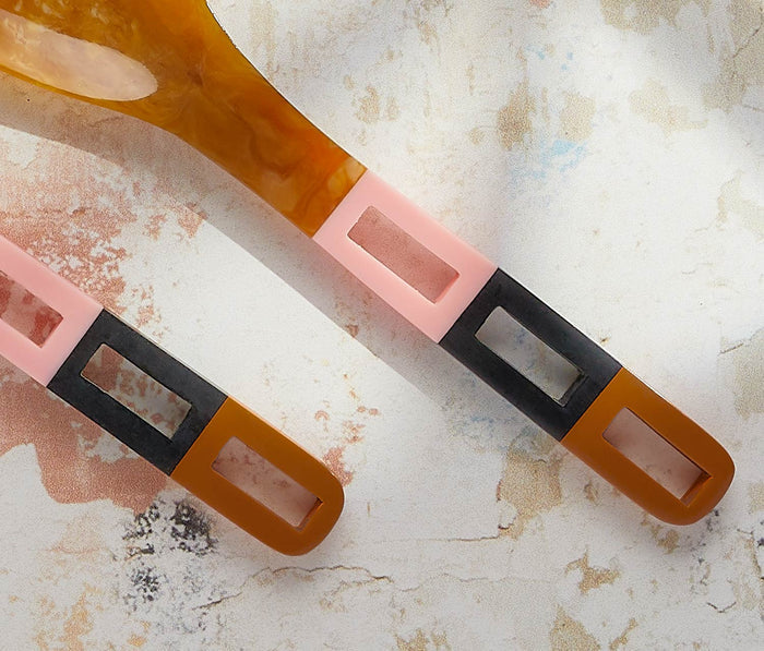 Two modern kitchen utensils with a translucent amber-brown handle, a light pink middle section, and a black rectangular accent, resting on a textured off-white surface.