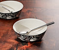 Two black ceramic Swan Song Imprint Cereal side bowls with white swan and floral patterns, shown on a dark wooden table with silver spoons inside.