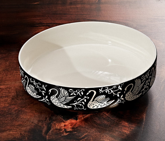 A large round serving bowl stoneware with a glossy white interior and a black exterior decorated with a white swan and floral pattern, shown on a dark wooden surface- Lollygag
