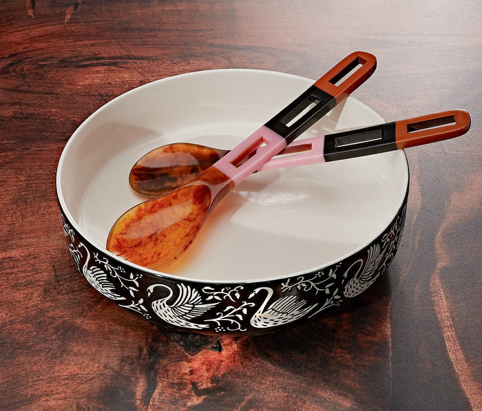 A white ceramic bowl with a black swan pattern around the rim, holding two tortoiseshell spoons with pink and black handles, set on a dark wooden table.