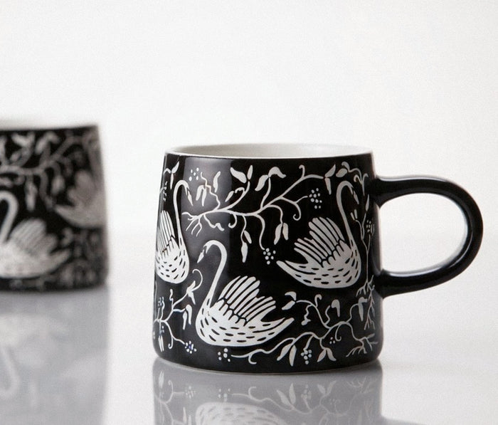 A black ceramic mug with a white hand-painted style swan and floral pattern, shown on a reflective white surface- Lollygag