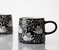 A black ceramic mug with a white hand-painted style swan and floral pattern, shown on a reflective white surface- Lollygag