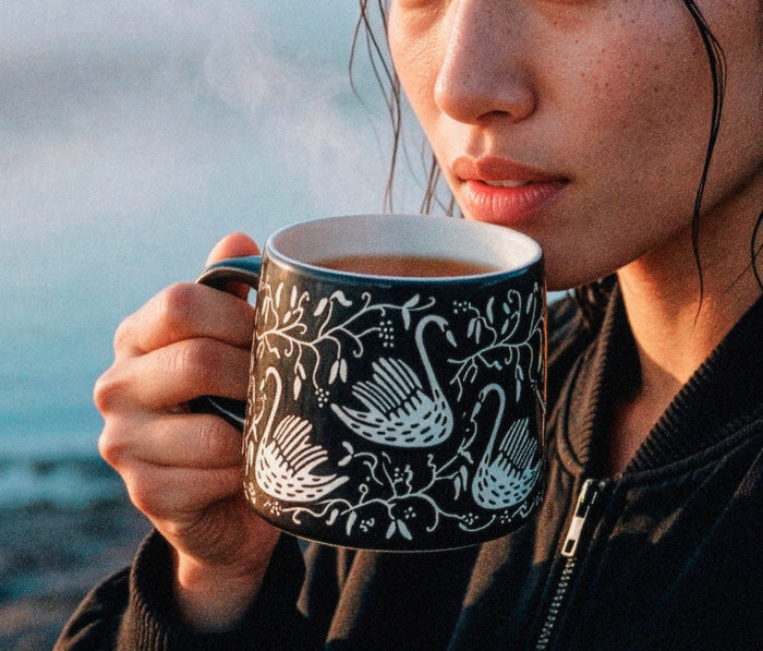 A close-up lifestyle shot of a person holding a black ceramic mug with a white swan and floral pattern, with steam rising from the tea inside, set against a soft-focus outdoor background - Lollygag