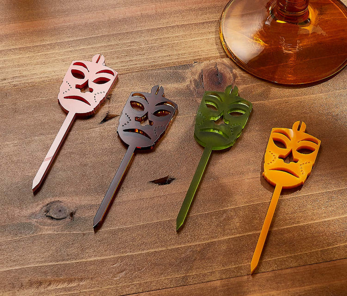 Four mask-shaped cocktail stirrers on a wooden surface with a glass of amber liquid in the background.