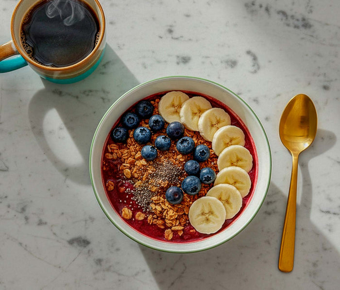 A bowl of acai bowl with bananas and blueberries, accompanied by a cup of coffee on a marble surface.